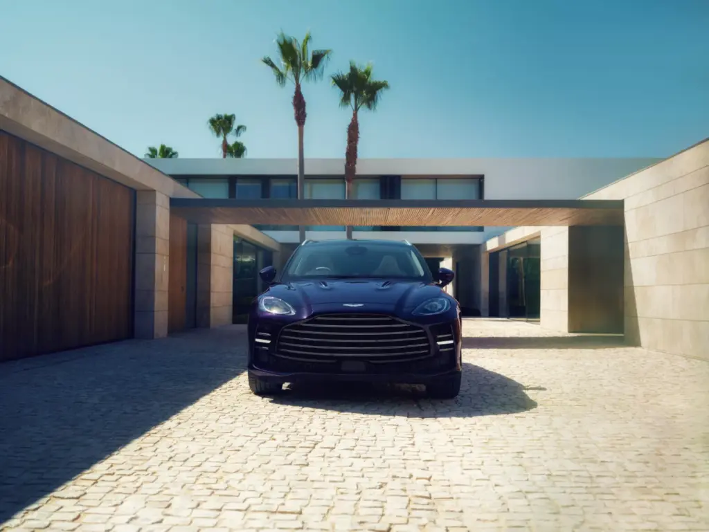 A dark blue Aston Martin DBX GT parked in a driveway outside a modern house with palm trees.