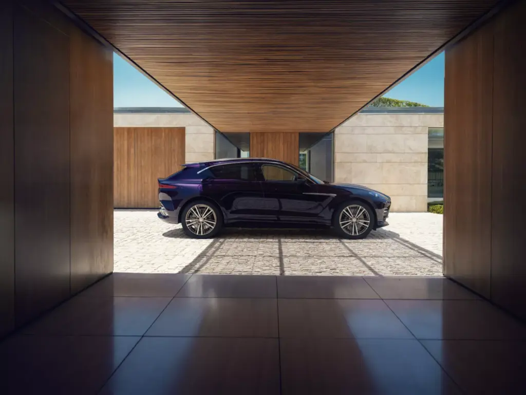 A dark purple Aston Martin DBX GT parked under a wooden canopy in front of a modern stone house.