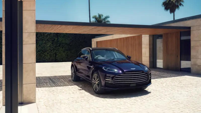 A dark purple Aston Martin DBX GT parked under a modern carport in front of a luxury villa with palm trees.