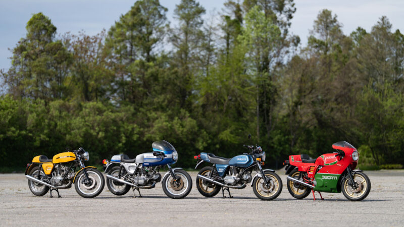 Four vintage Ducati motorcycles in yellow, silver, blue, and red parked in a row against a tree line.