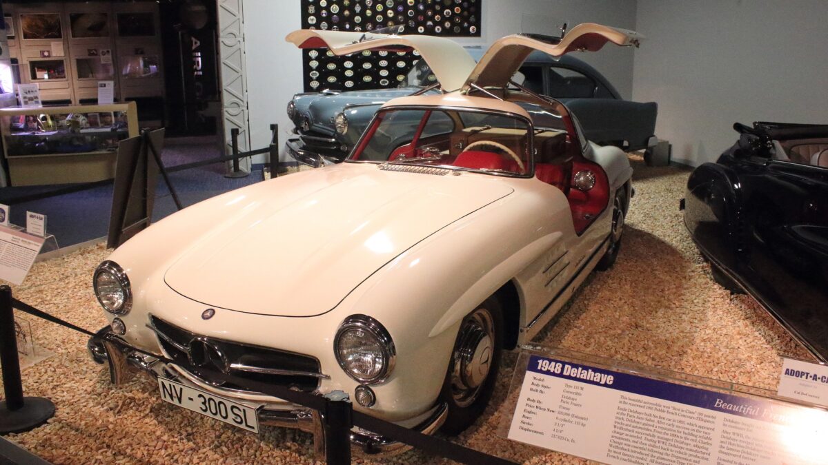 A white Mercedes-Benz 300 SL Gullwing with its doors raised, displayed in a museum among other classic cars.