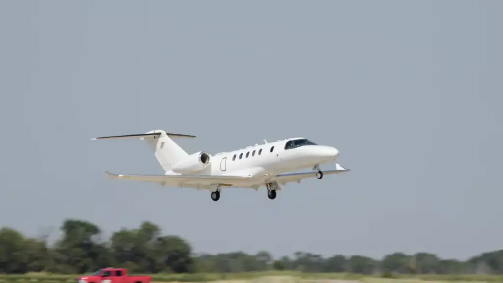 A white business jet climbs into the sky just after takeoff from a runway.