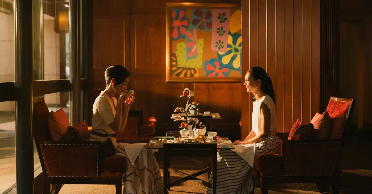 Two women sit at a table having afternoon tea in an elegant hotel lounge with warm lighting.
