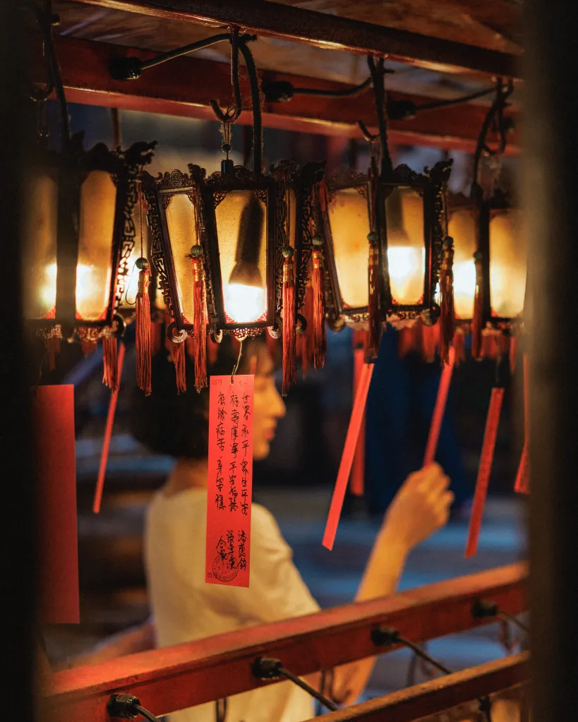 Traditional Chinese lanterns with red tasseled tags hang in a temple, with a person visible in the background.