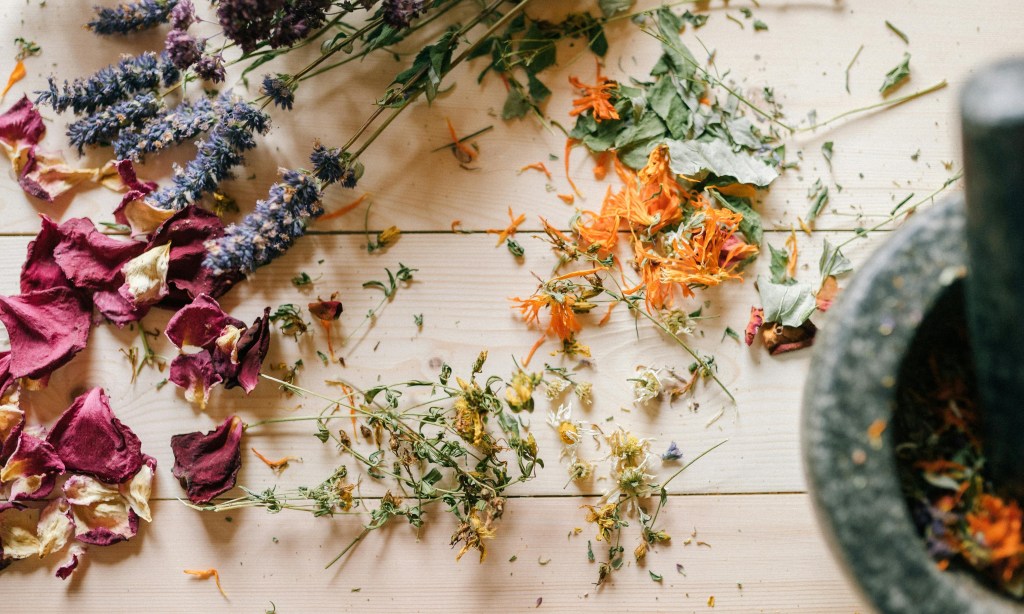 Dried flower petals and herbs scattered on a wooden surface beside a stone mortar and pestle.