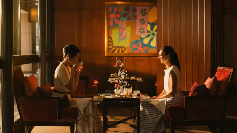 Two women sit at a table having afternoon tea in an elegant hotel lounge with warm lighting.