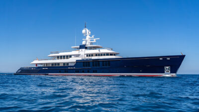 A dark blue superyacht with a white superstructure sails on calm blue water under clear skies.