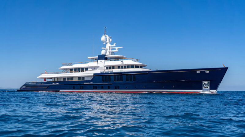 A dark blue superyacht with a white superstructure sails on calm blue water under clear skies.
