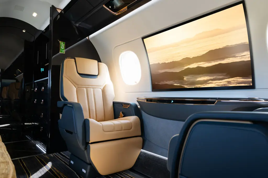 Interior of a private jet cabin with a tan leather seat and a large screen showing a mountain sunset view.