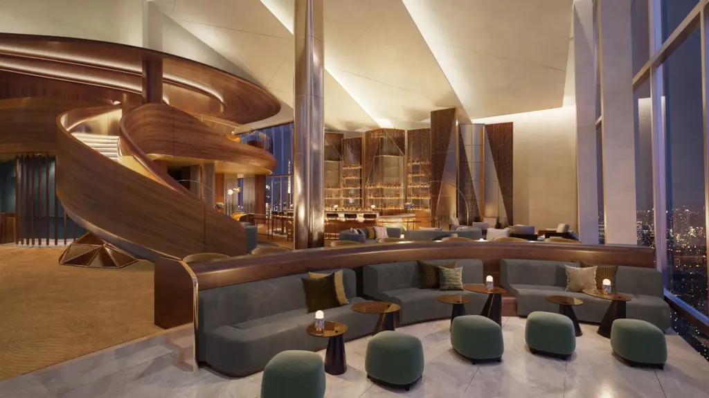 A modern hotel lounge with a curved wooden staircase, gray sofas, green ottomans, and a night city skyline view.