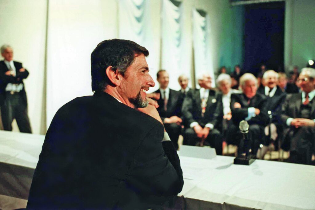 A man in a suit smiles in profile at a formal event with an audience seated behind him.