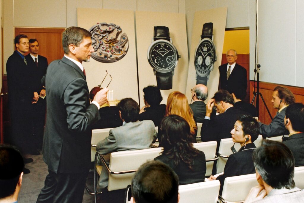 A man addresses seated attendees in front of posters showing A. Lange & Söhne watches and a movement.