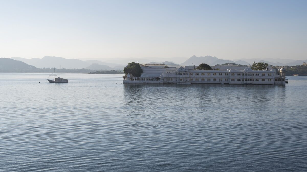 The white Taj Lake Palace hotel sits on Lake Pichola with mountains in the background and a boat nearby.