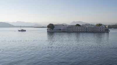 The white Taj Lake Palace hotel sits on Lake Pichola with mountains in the background and a boat nearby.