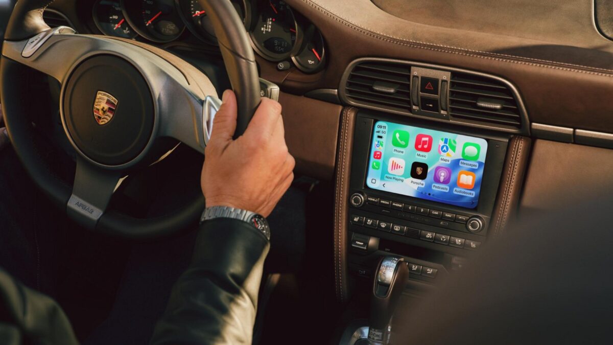 A hand on a Porsche steering wheel with the PCCM Plus touchscreen displaying CarPlay icons.