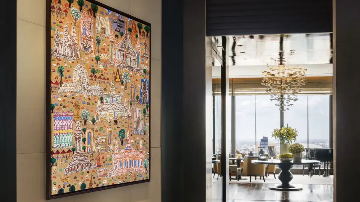 A large colorful painting of temple-like buildings hangs in a hotel corridor overlooking a lounge with city views.