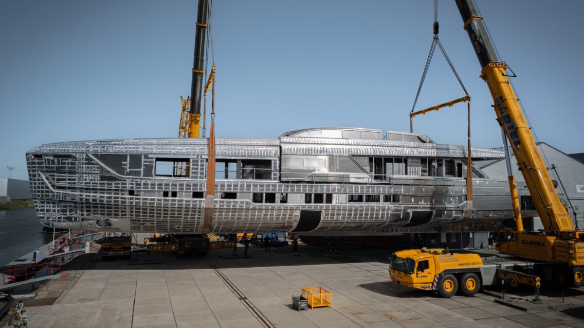 A large unpainted aluminum yacht hull and superstructure being lifted by two cranes at a shipyard.
