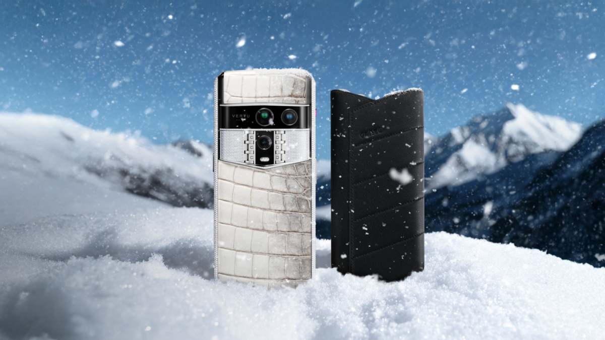 A Vertu smartphone with a white crocodile-textured back and a black leather case stand in snow with mountains behind.