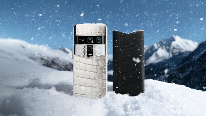 A Vertu smartphone with a white crocodile-textured back and a black leather case stand in snow with mountains behind.