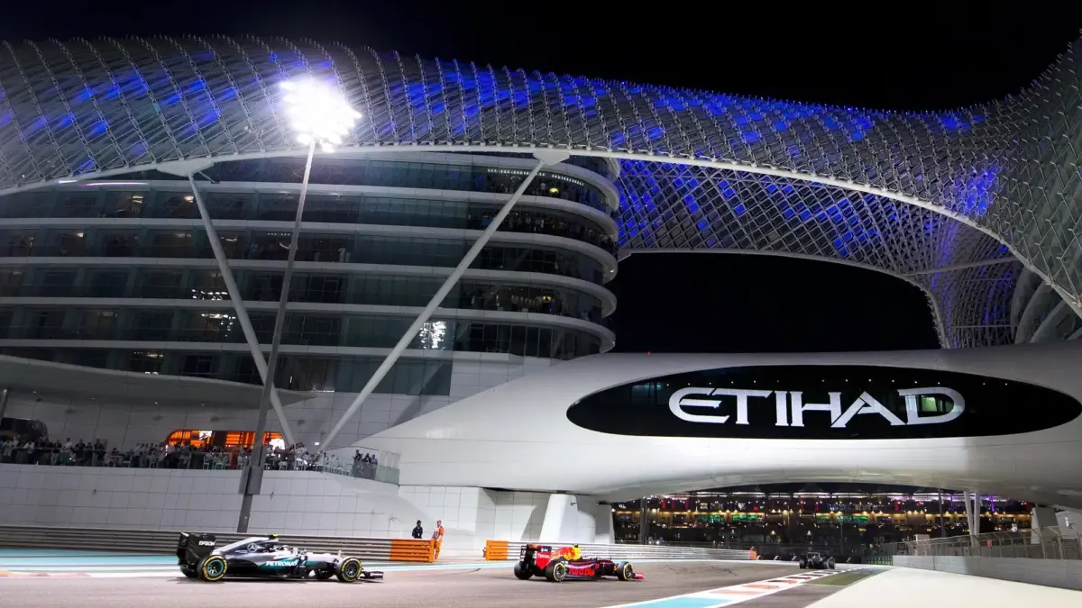 Formula 1 cars race at night under the Yas Marina Circuit hotel bridge with the Etihad logo.