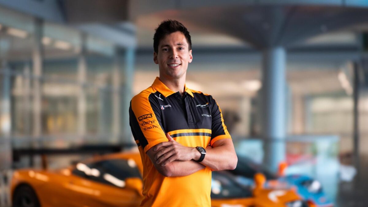 A man in orange and black McLaren racing team polo shirt stands with arms crossed in front of McLaren race cars.