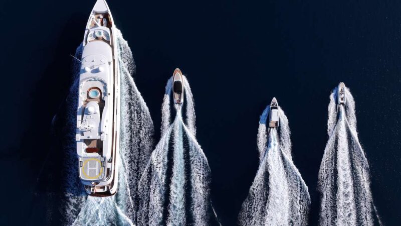 Aerial view of a large white superyacht trailed by three smaller boats, all leaving white wakes on dark blue water.