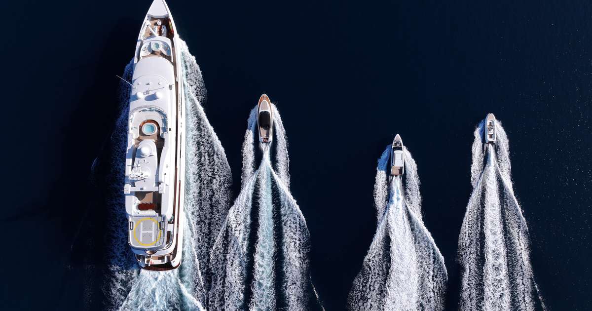 Aerial view of a large white superyacht trailed by three smaller boats, all leaving white wakes on dark blue water.
