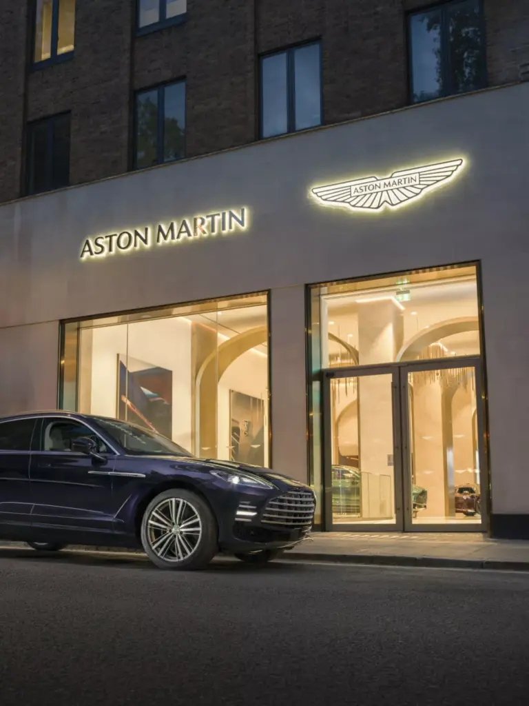 An Aston Martin showroom storefront at night with illuminated signage and a dark Aston Martin car parked outside.