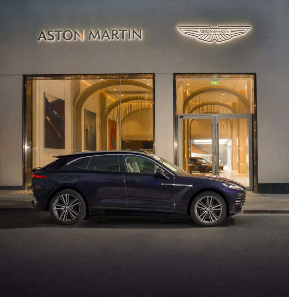 A dark Aston Martin SUV parked outside an illuminated Aston Martin showroom at night.