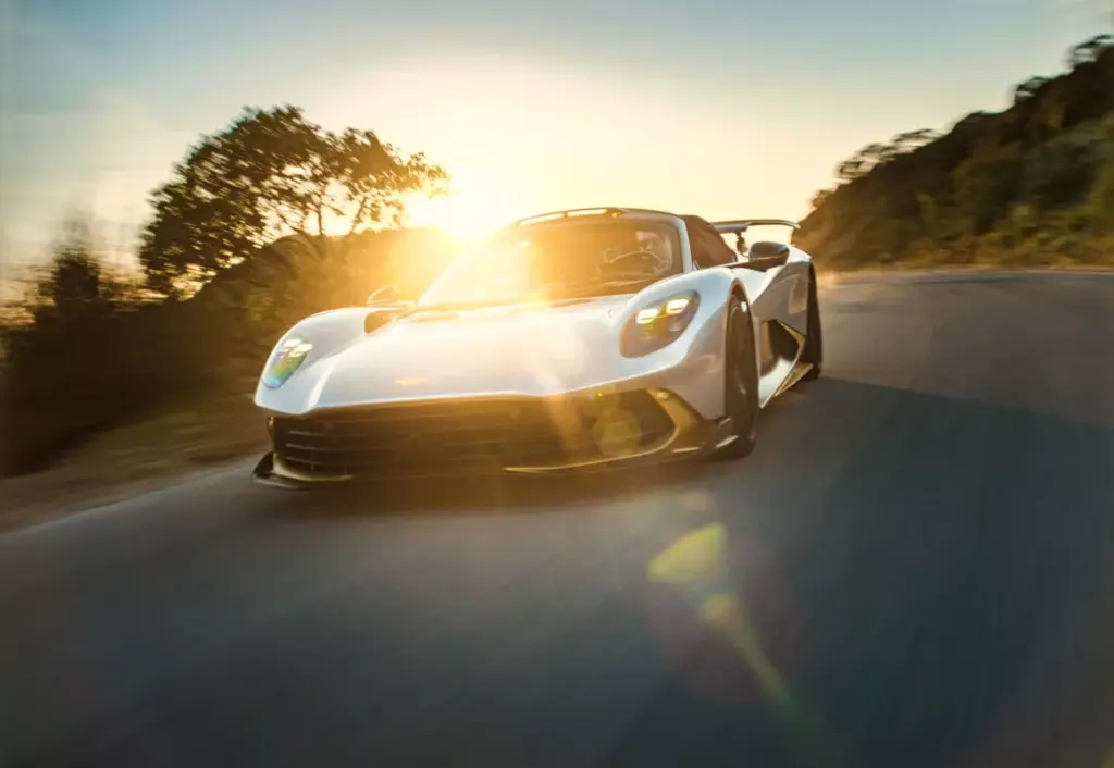 White Aston Martin Valhalla Spider sports car driving on a road at sunset with sun flare.