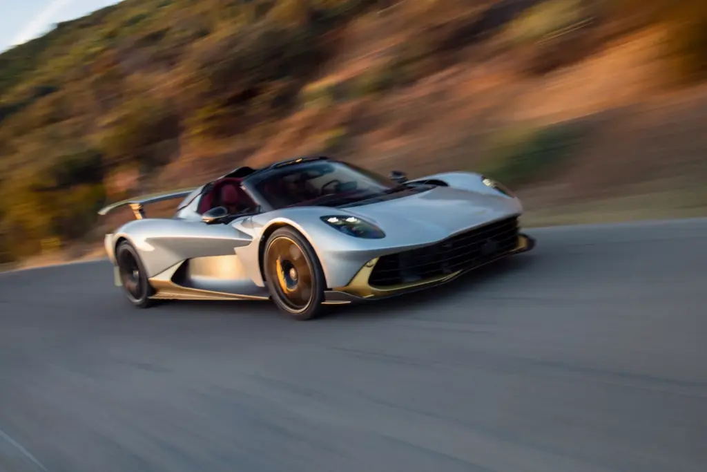 A silver and gold Aston Martin Valhalla Spider speeds down a mountain road with its roof removed.