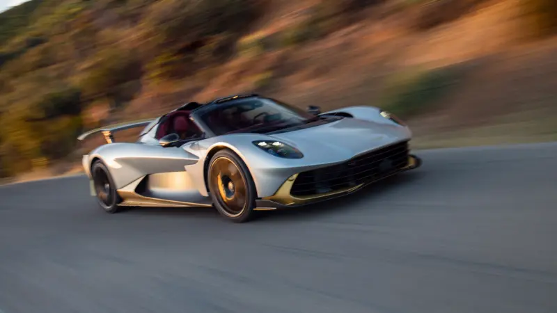 A silver and gold Aston Martin Valhalla Spider speeds along a mountain road with motion blur in the background.