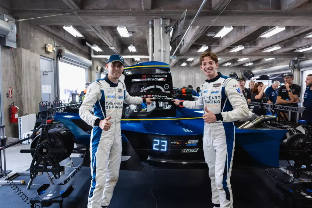 Two racing drivers in white suits give thumbs up in front of a blue Aston Martin Valkyrie race car in a garage.