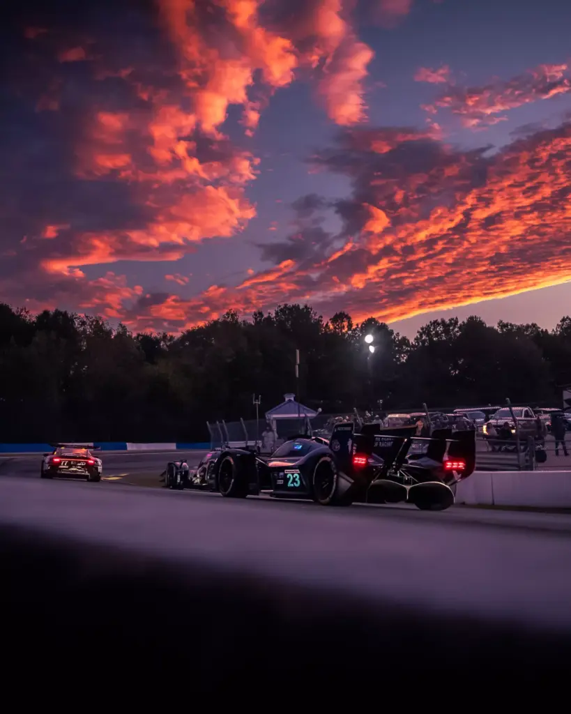 A race car numbered 23 on a racetrack at dusk under a vivid orange and purple cloudy sky.