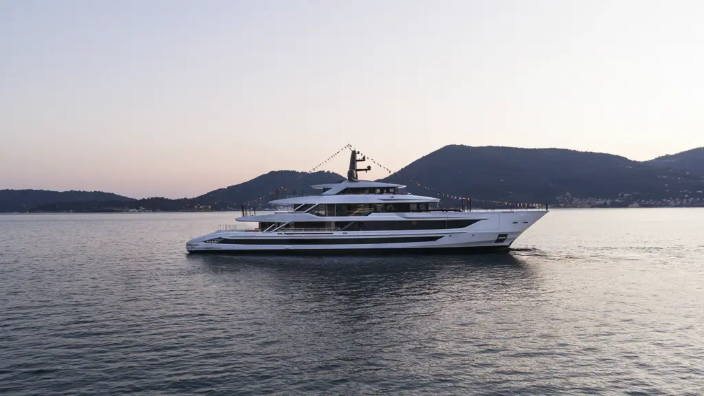 A large white superyacht decorated with signal flags floats on calm water with hills in the background at dusk.