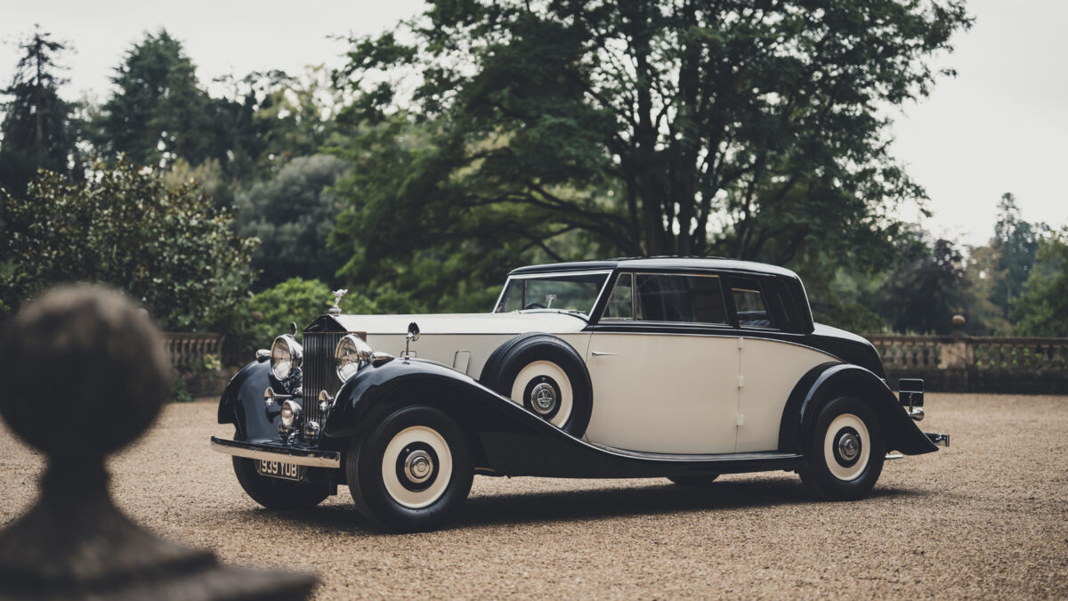 A black and cream vintage Rolls-Royce parked on a gravel driveway with trees in the background.