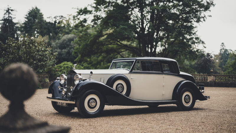 A black and cream vintage Rolls-Royce parked on a gravel driveway with trees in the background.