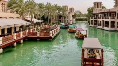 Wooden abra boats float on a green canal beside traditional Arabian-style buildings and palm trees at Madinat Jumeirah.