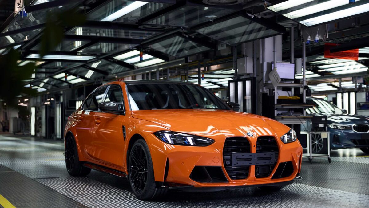 An orange BMW M3 sedan sits on a factory production line surrounded by assembly equipment.