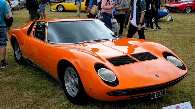 An orange Lamborghini Miura parked on grass at a car show with spectators in the background.