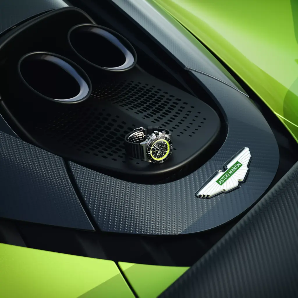 A watch rests on the carbon-fiber bodywork of a green Aston Martin near the exhaust and Aston Martin badge.