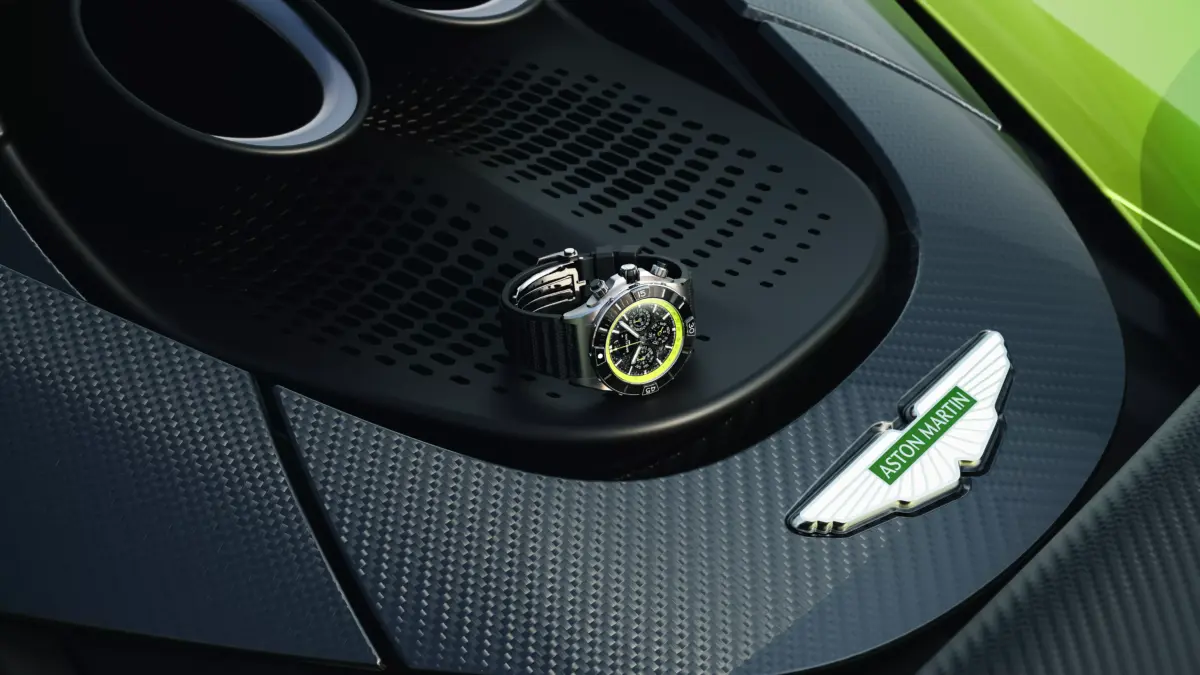 A watch rests on the carbon-fiber bodywork of a green Aston Martin near the exhaust and Aston Martin badge.