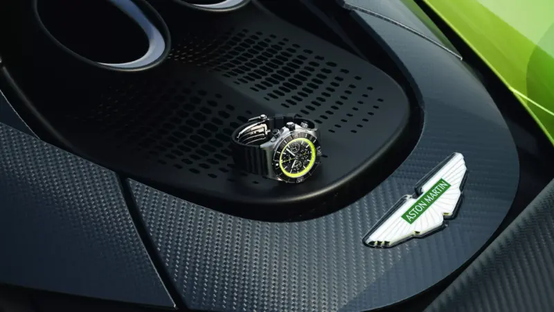 A watch rests on the carbon-fiber bodywork of a green Aston Martin near the exhaust and Aston Martin badge.