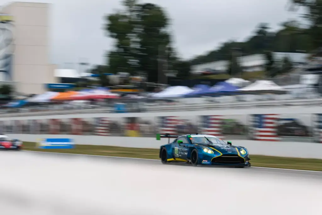 A blue Aston Martin Vantage GT3 race car speeds along a track with blurred grandstands in the background.