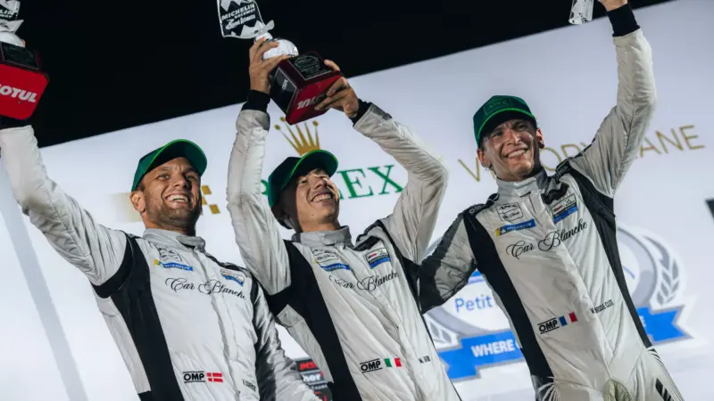 Three racing drivers in Car Blanche team suits celebrate on a podium holding trophies.