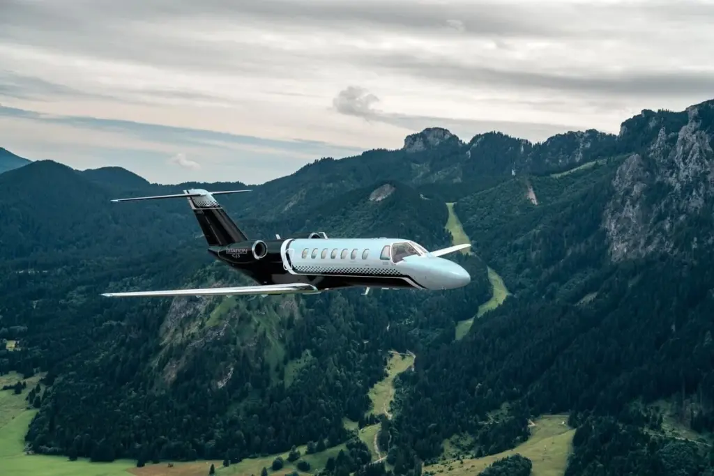 A black and white Cessna Citation CJ3 business jet flying over forested mountains.
