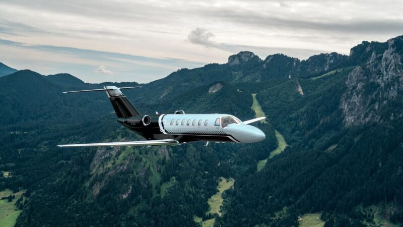 A Cessna Citation CJ3 business jet flying over forested mountains.