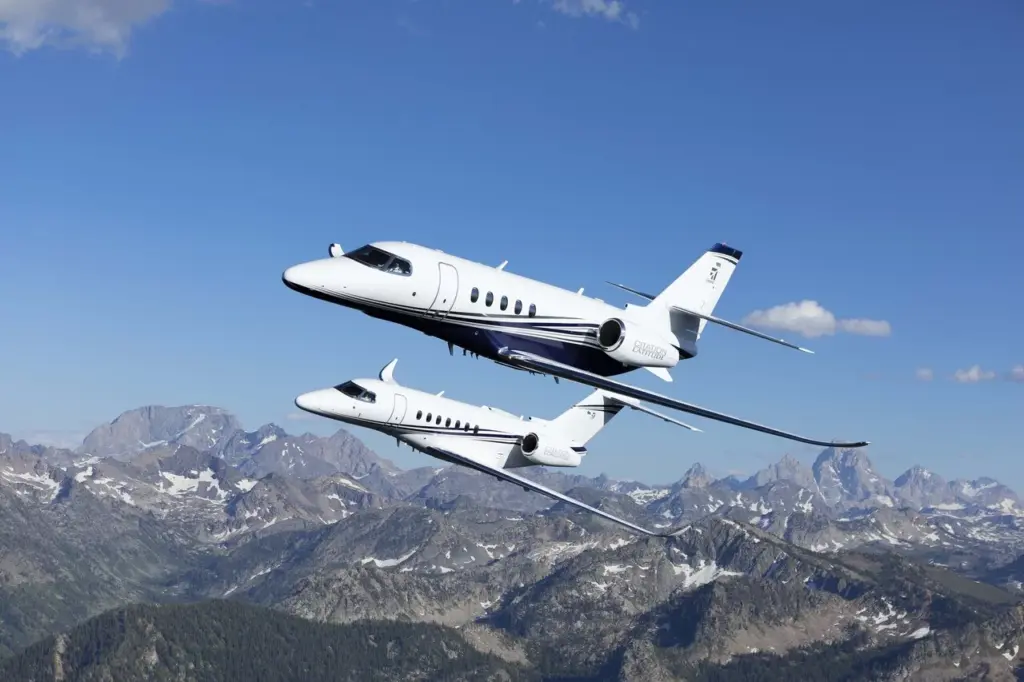 Two Cessna Citation business jets fly in formation over a mountain range under a blue sky.