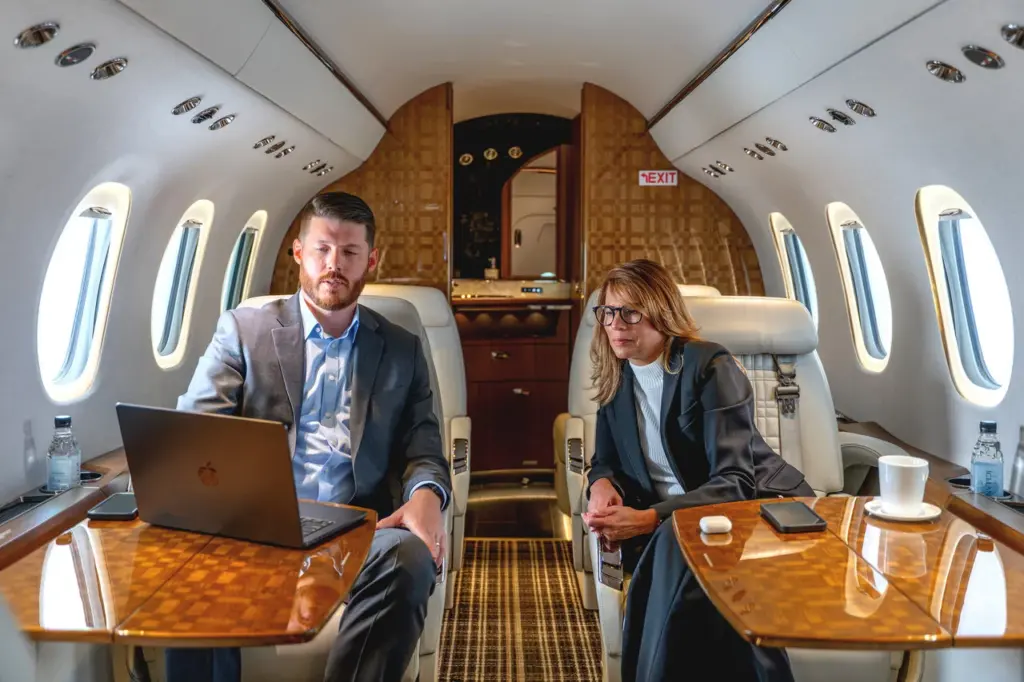 Two passengers sit at wood tables aboard a private jet cabin, one using a laptop.