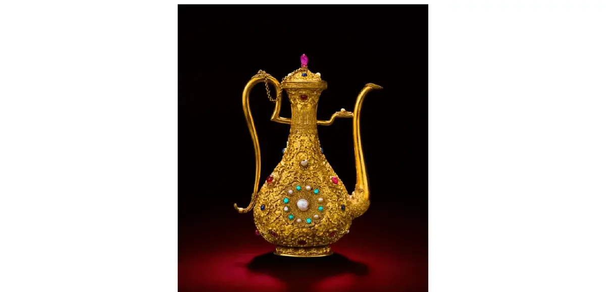 A gold ewer encrusted with gemstones and pearls, photographed against a dark red background.